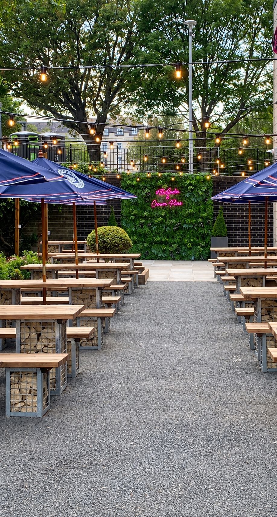 An outdoor seating area with tables and umbrellas, a gravel path, and a hedge with a neon sign.