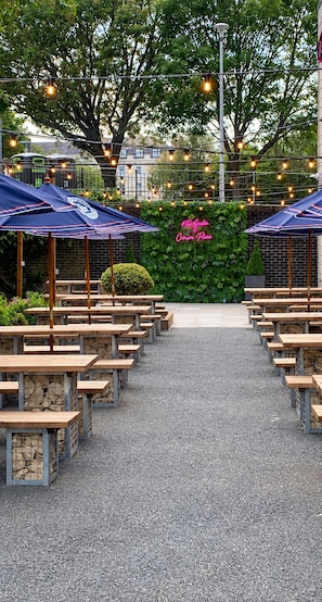 An outdoor seating area with tables and umbrellas, a gravel path, and a hedge with a neon sign. - Crowne Plaza Plymouth by IHG (Plymouth)