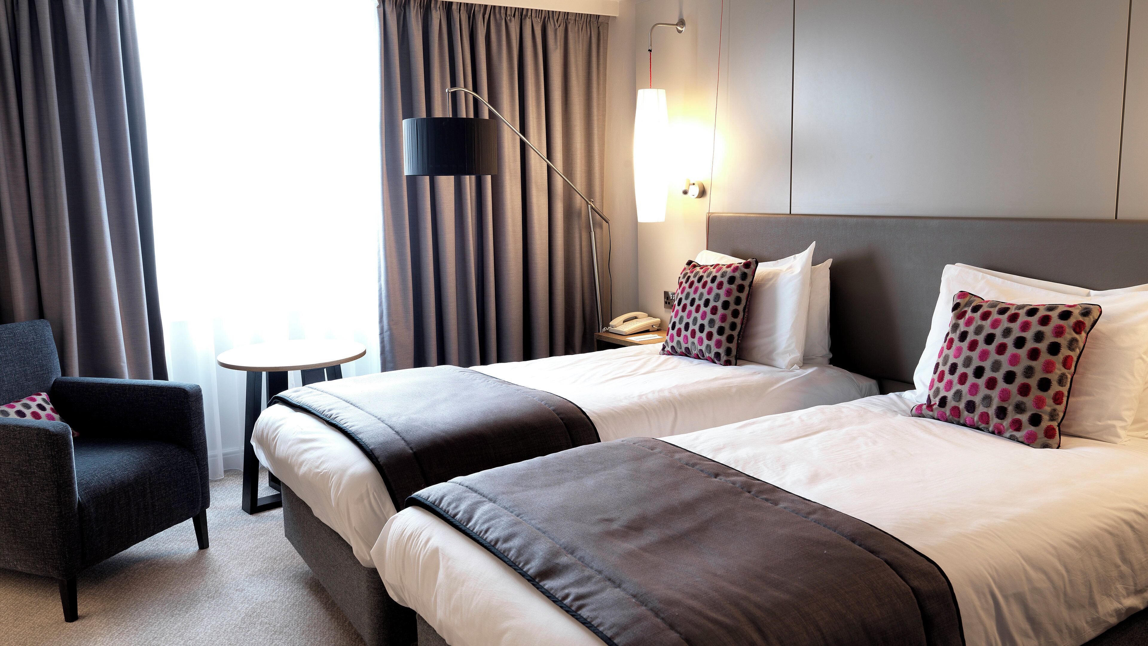 Quarto de hotel com duas camas, uma cadeira, mesa de cabeceira com abajur e vista para o exterior.