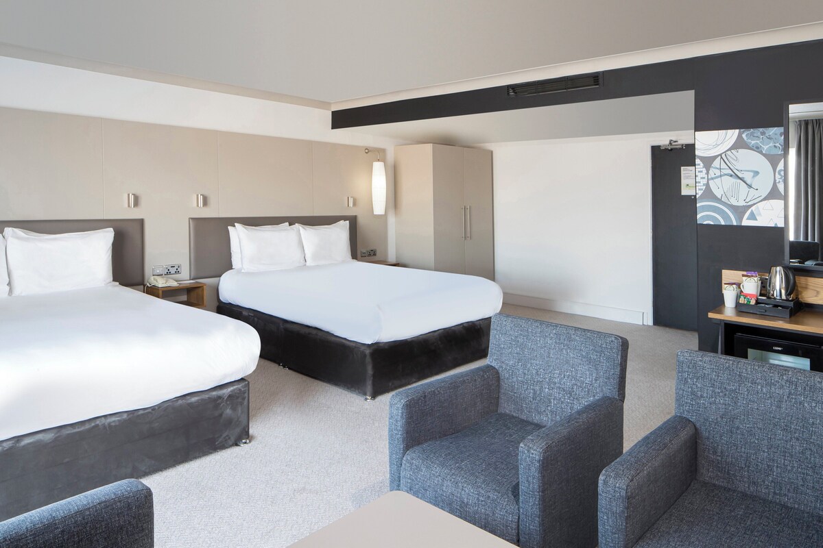 A hotel room with two beds, a desk, a chair, and a small table with a kettle.
