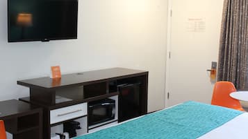 A hotel room with a bed, a desk with a microwave, a TV, and a door with an emergency evacuation sign.
