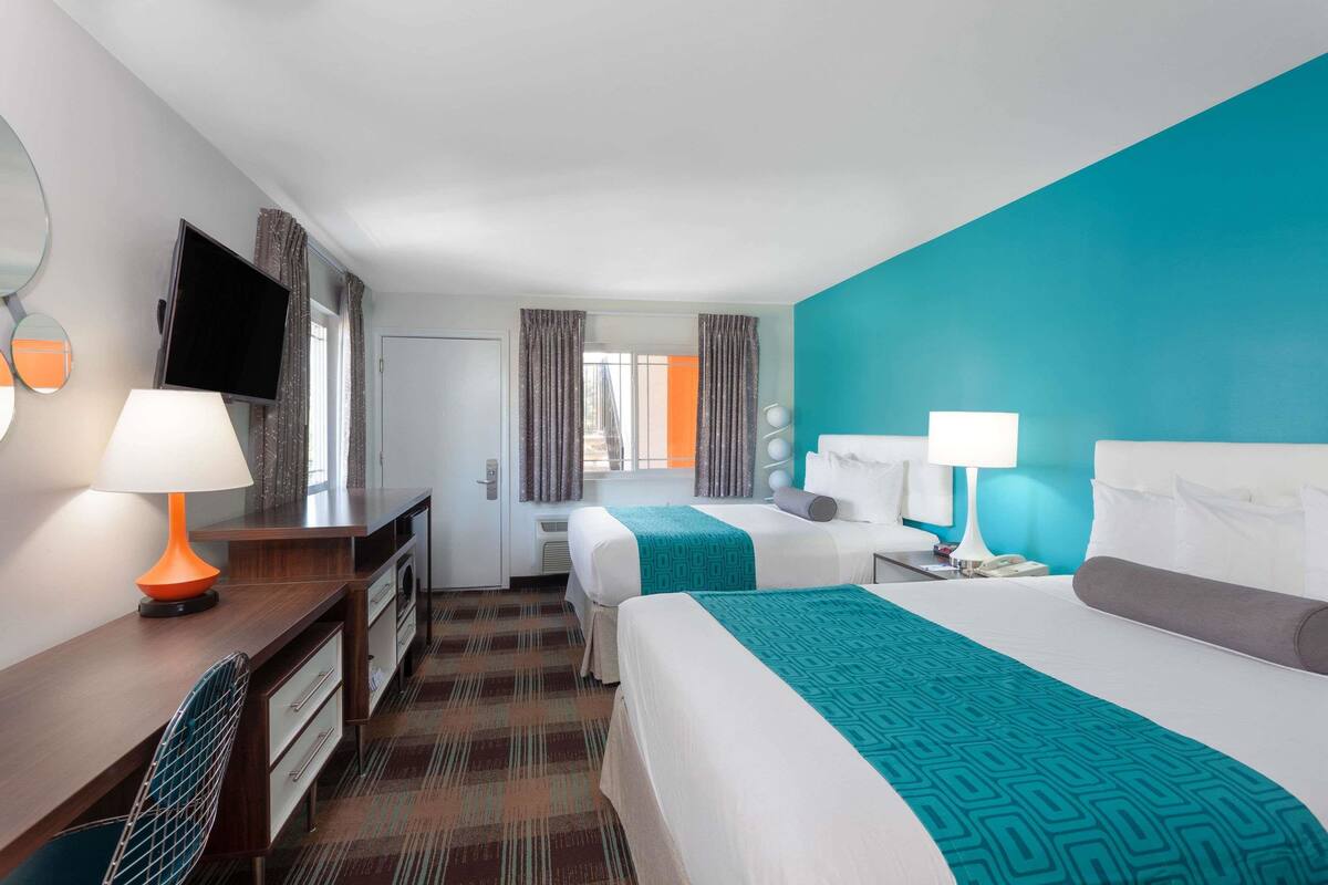 Habitación de hotel con dos camas, un escritorio con televisor, una silla y un baño visible a través de la ventana.