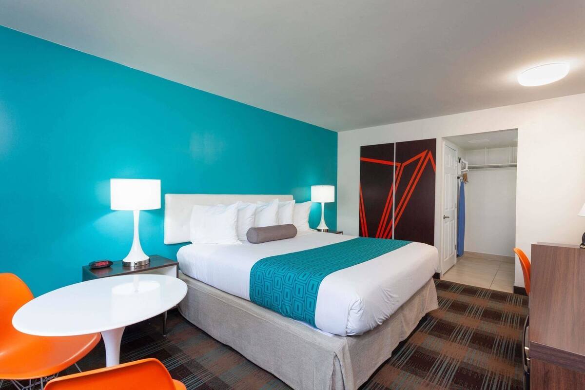 Una habitación de hotel con una cama, dos mesitas de noche con lámparas, una pequeña mesa redonda con dos sillas naranjas, un armario y un espejo.