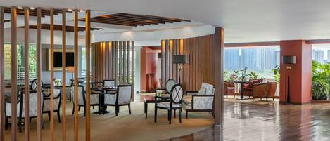 A spacious lobby with wooden flooring, a seating area with chairs and tables, and a view of tall buildings.