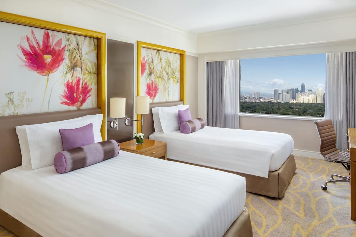 Habitación de hotel con dos camas, un escritorio y una silla. Muebles con grandes cuadros florales en las paredes y vista a un paisaje urbano por la ventana.