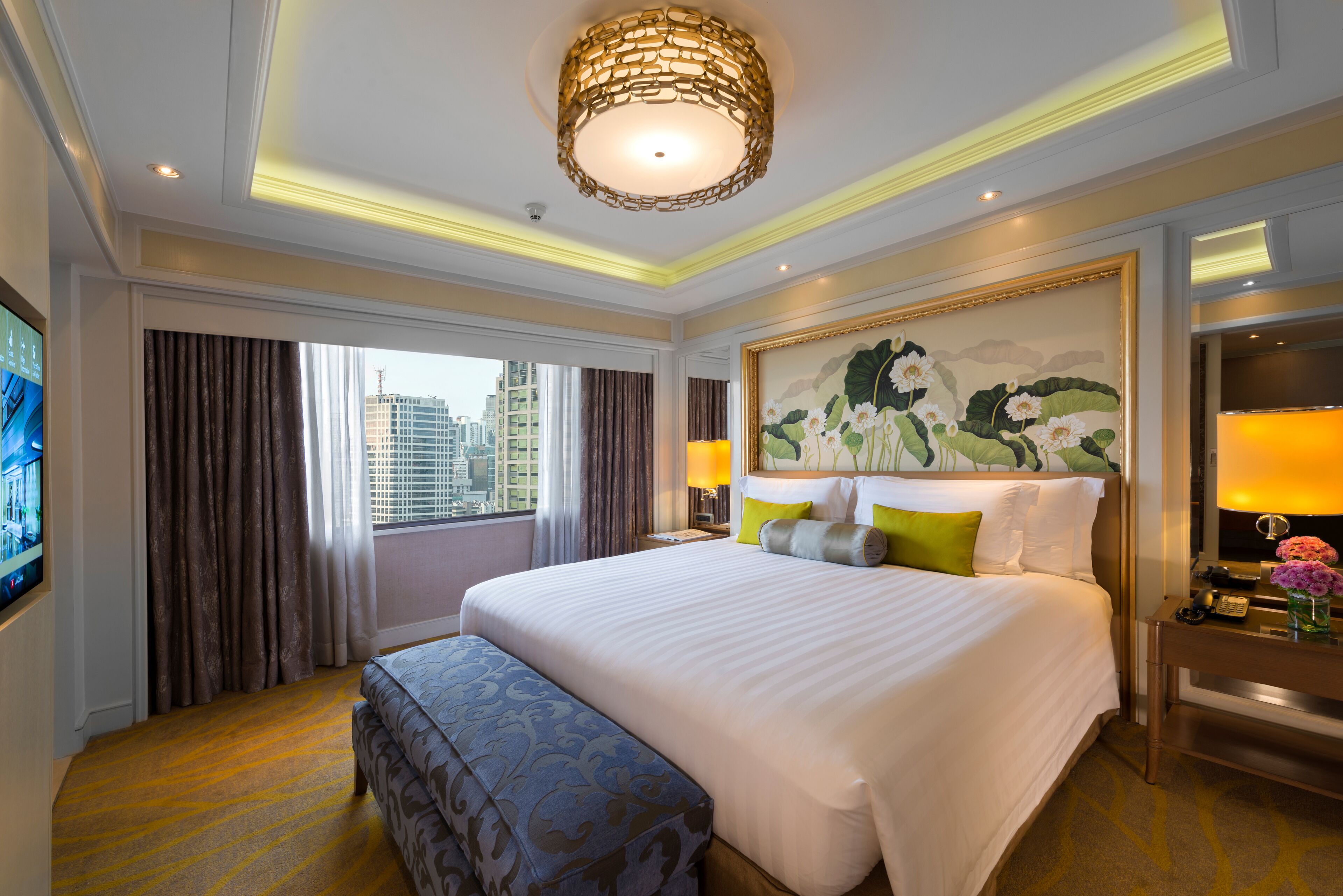 A hotel room with a large bed, a desk with a lamp, a painting on the wall, and a view of the city through the window.