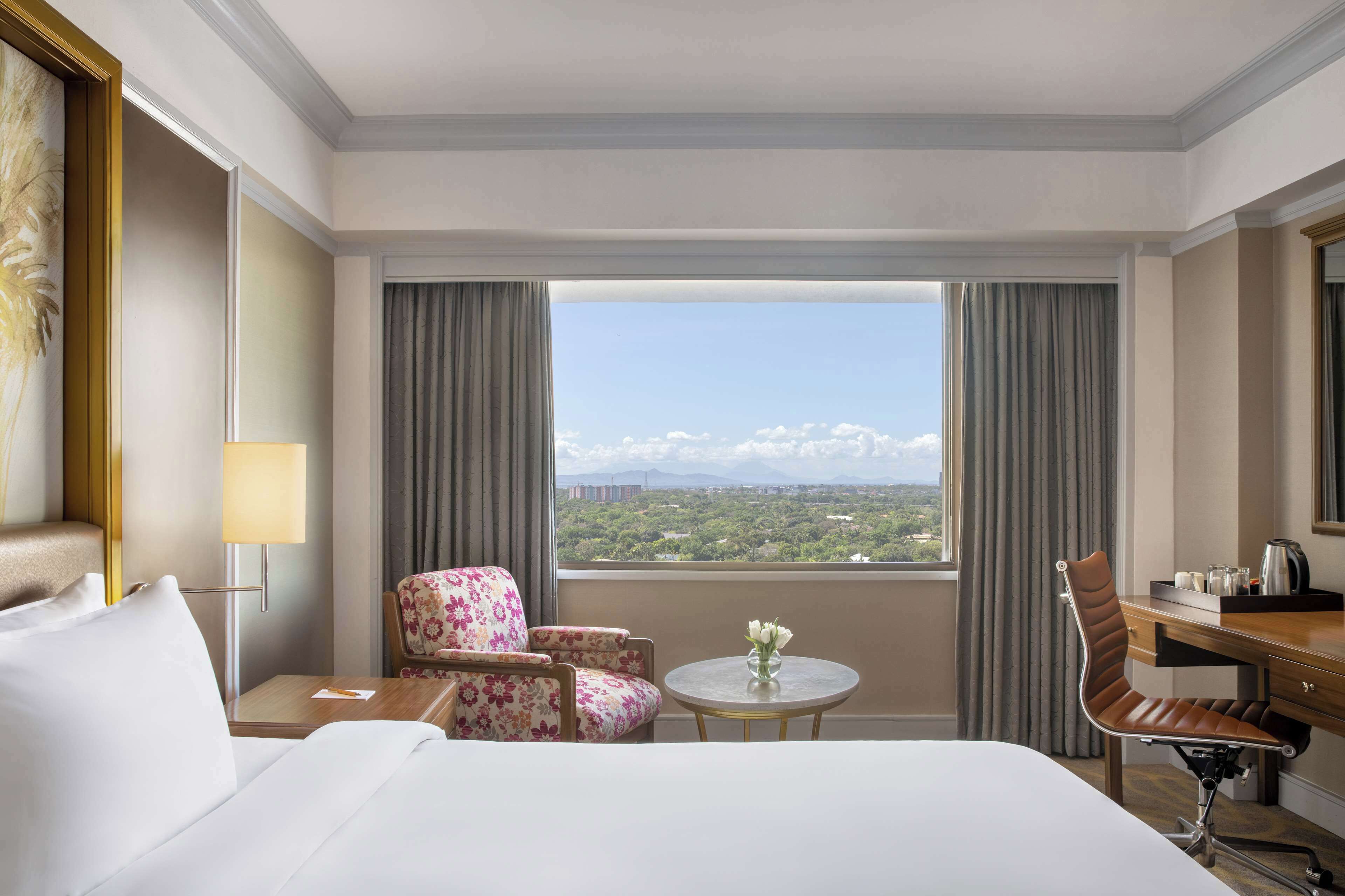 A hotel room with a bed, two floral armchairs, a desk with a chair, a small table with a vase, and a view of a mountainous landscape through large windows.