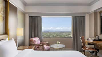 A hotel room with a bed, two floral armchairs, a desk with a chair, a small table with a vase, and a view of a mountainous landscape through large windows.