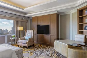 A hotel room with a large window, a flat-screen TV, a patterned carpet, a sofa, a chair, and a small side table with a lamp.