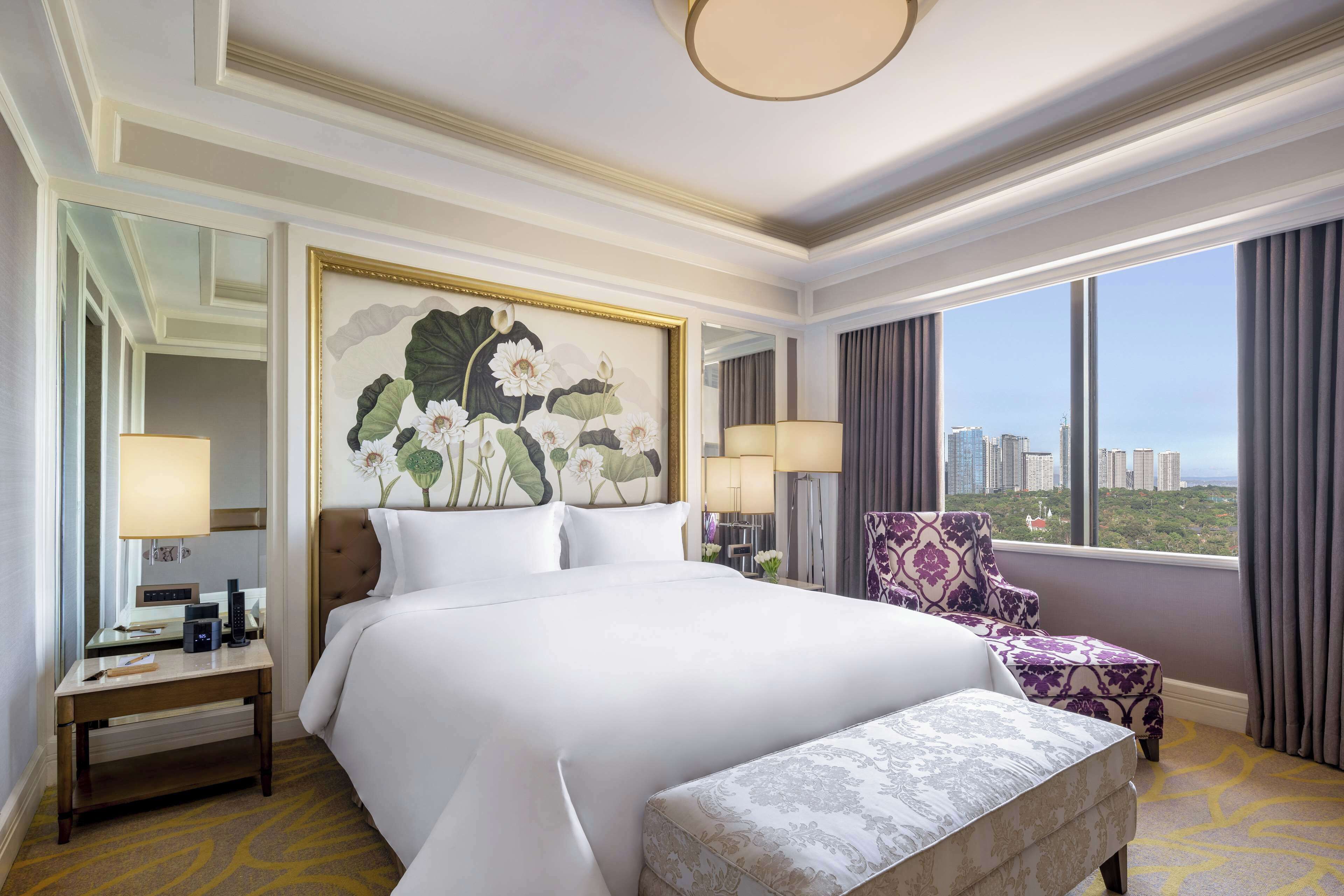 A hotel room with a large bed, a chair, a desk with a lamp, and a view of the city.