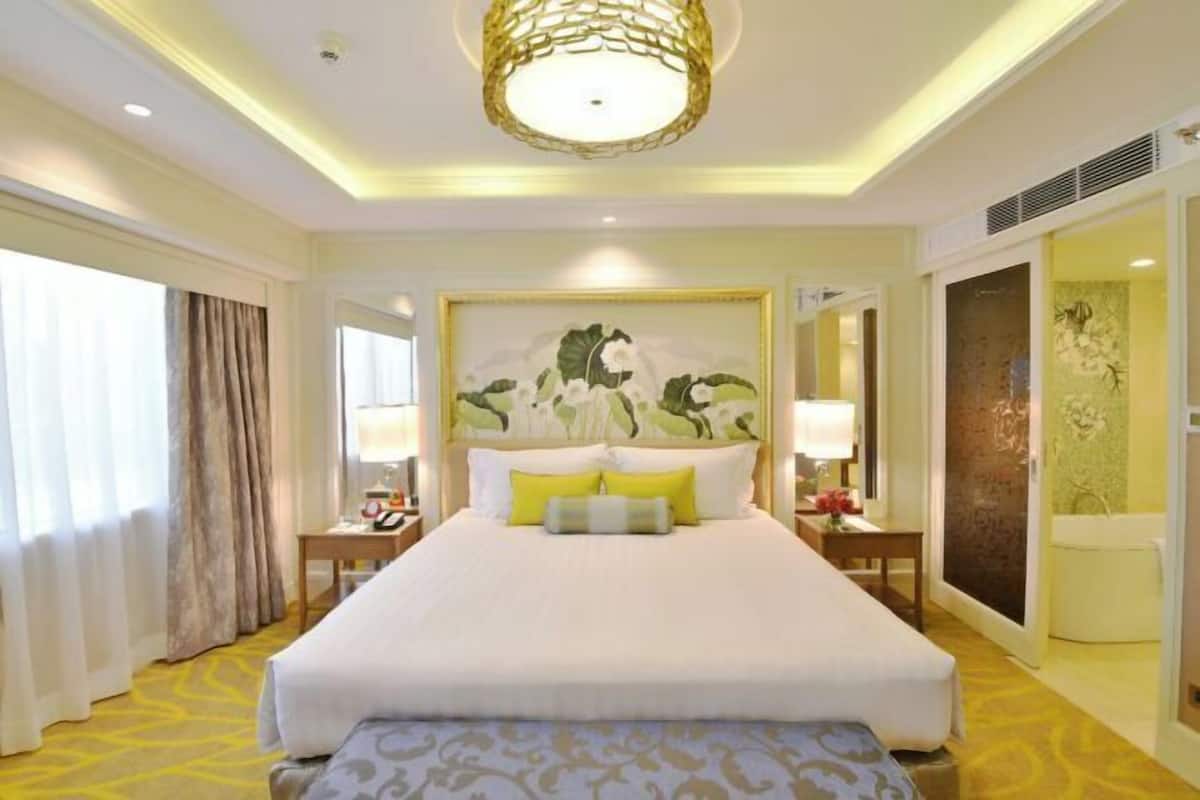 Habitación de hotel con una cama grande, mesitas de noche, cabecero con obra de arte, ventana con cortinas y baño a la vista a través de una puerta abierta.