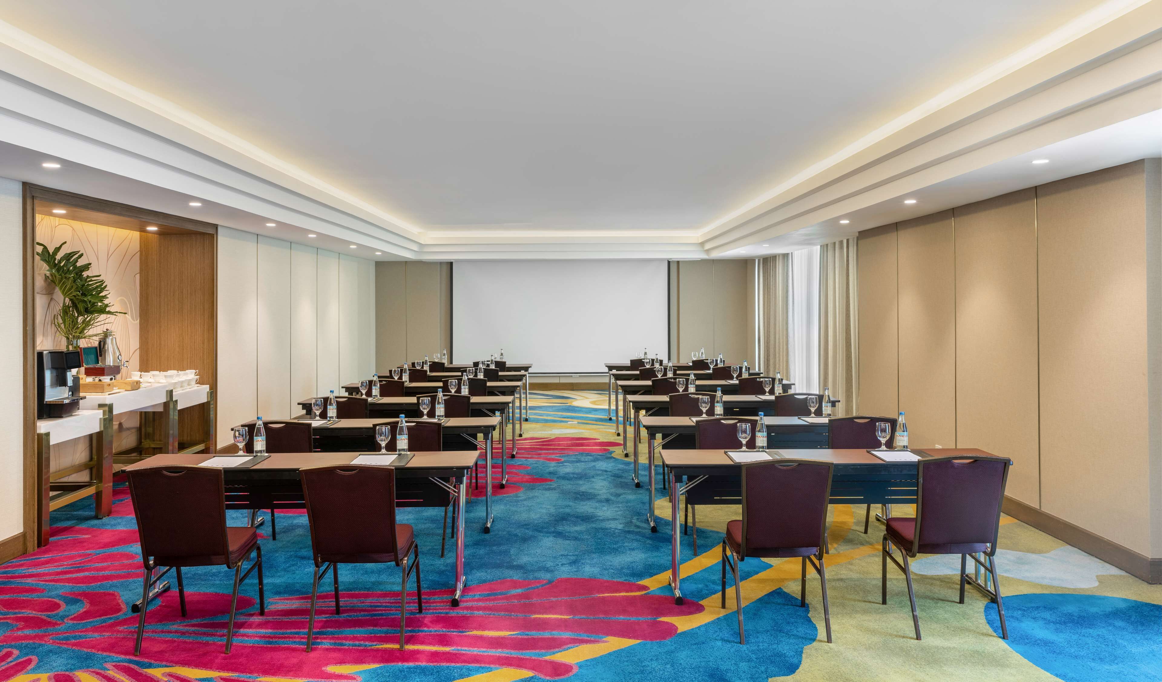 A conference room with rows of tables and chairs, a projector screen, and a carpeted floor.