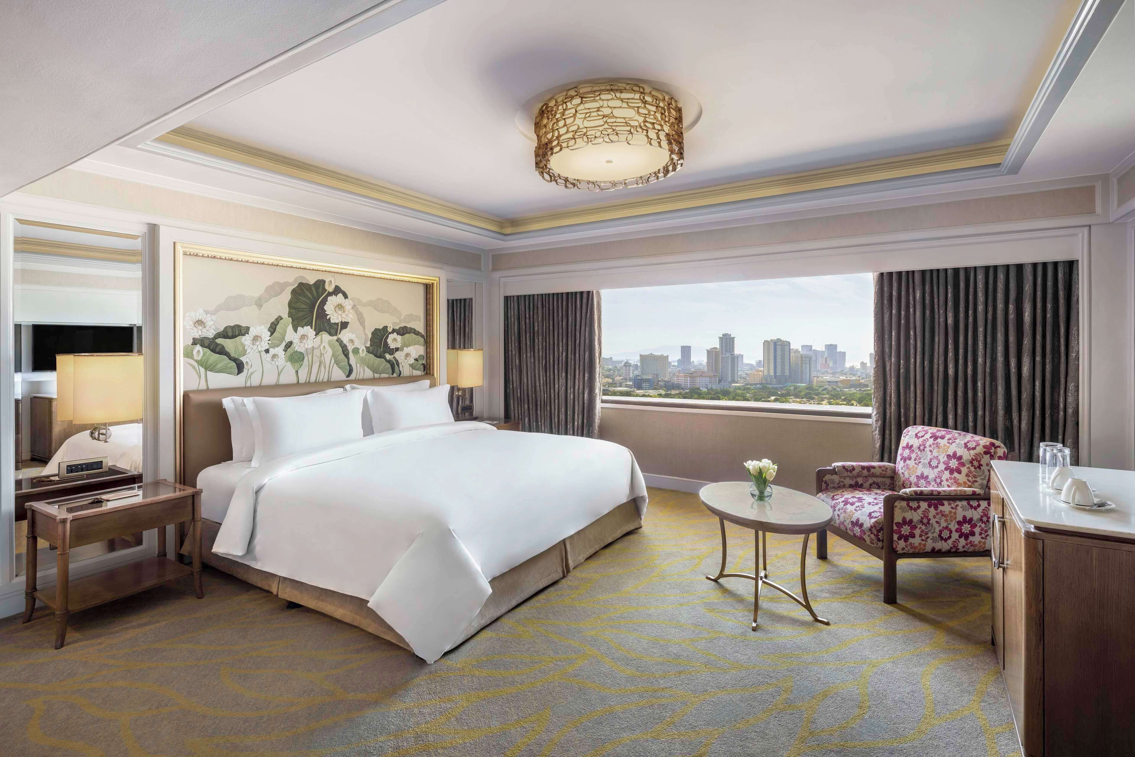 A hotel room with a large bed, a desk, a chair, a small table with a vase of flowers, and a city view through the window.
