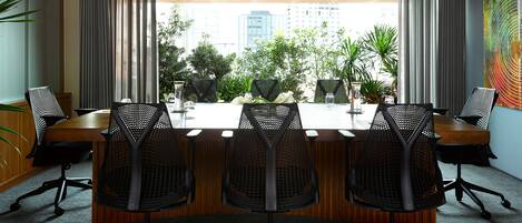 A conference room with a long table, four chairs, and a view of a cityscape through large windows.