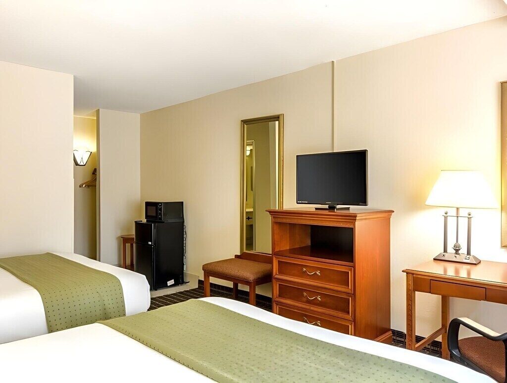 Ein Hotelzimmer mit zwei Betten, einem Fernseher, einem Schreibtisch mit Lampe und einem Spiegel.