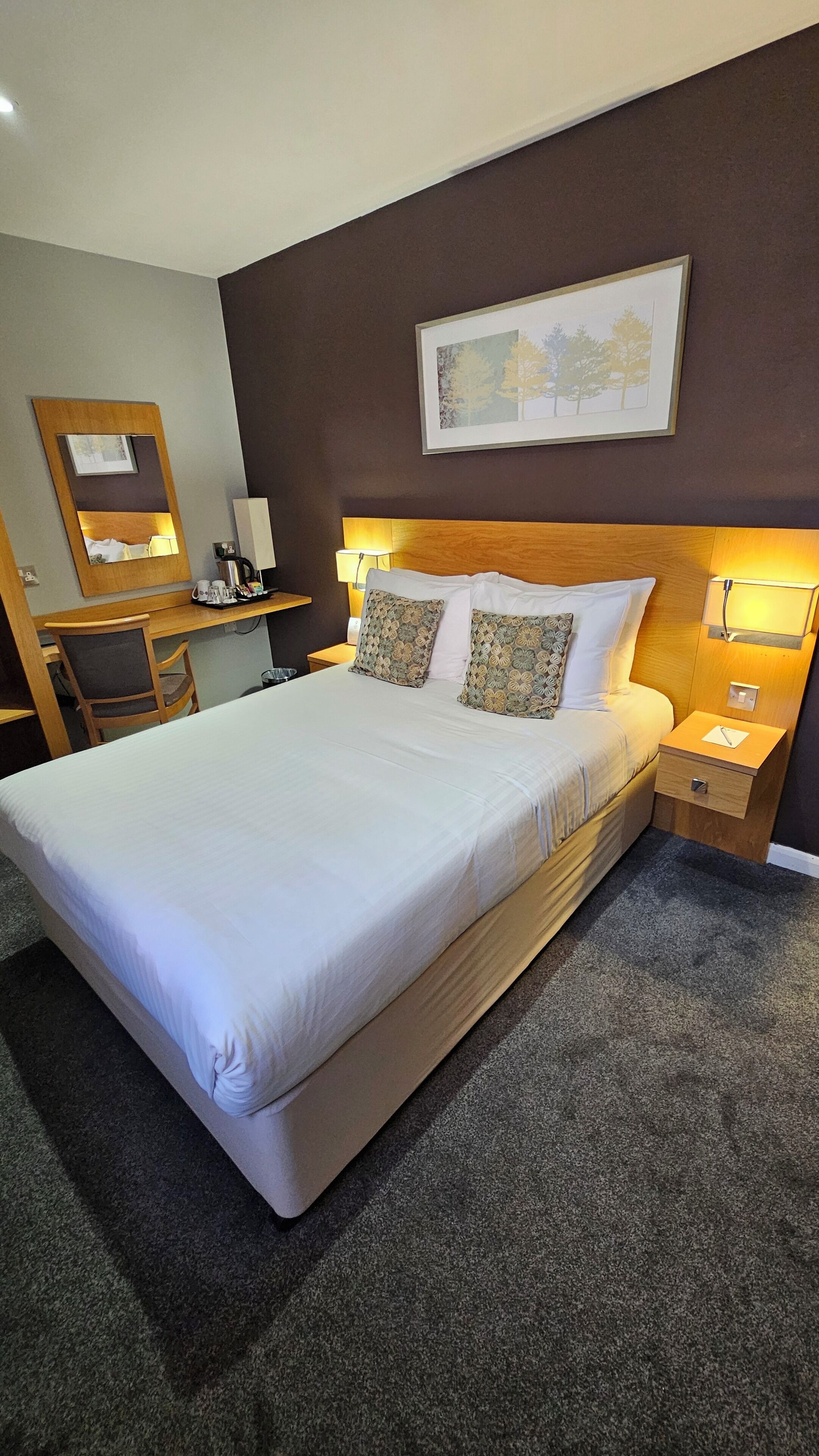Una habitación de hotel con una cama grande, un escritorio, una silla, una mesita de noche y una lámpara de pared.