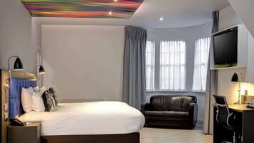 A modern hotel room with a large bed, a leather sofa, a desk with a computer, and a flat-screen TV.