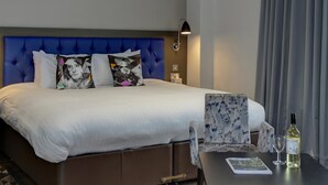 A modern hotel room with a tufted blue headboard, a bed with white linens, a small table with a bottle and glass, and a ceiling with a geometric light fixture. - Best Western Plus Nottingham Westminster Hotel (Nottingham)