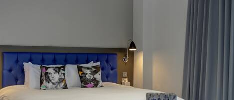 Une chambre d’hôtel moderne avec une tête de lit bleue à motifs capitonnés, un lit avec du linge de lit blanc, une petite table avec une bouteille et un verre, et un plafond agrémenté d’un luminaire aux formes géométriques.