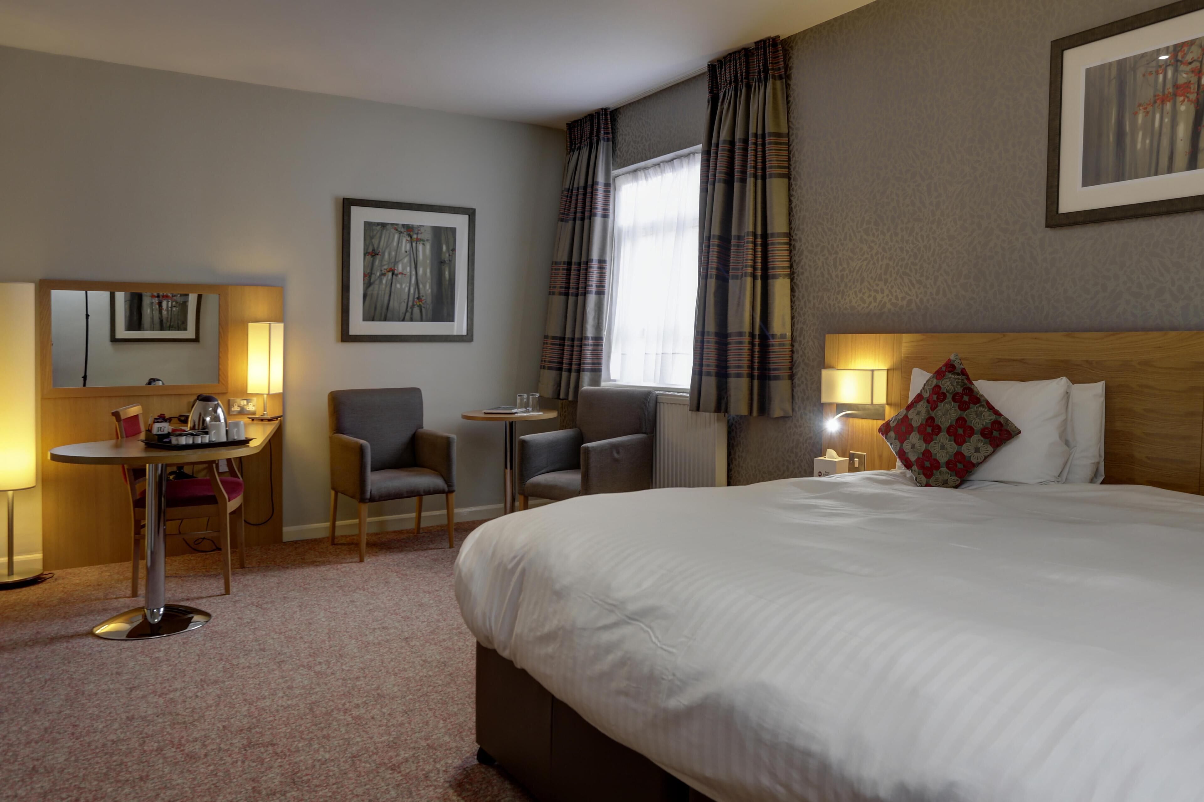 A hotel room with a large bed, a desk with a chair, a small table, and a window with curtains.
