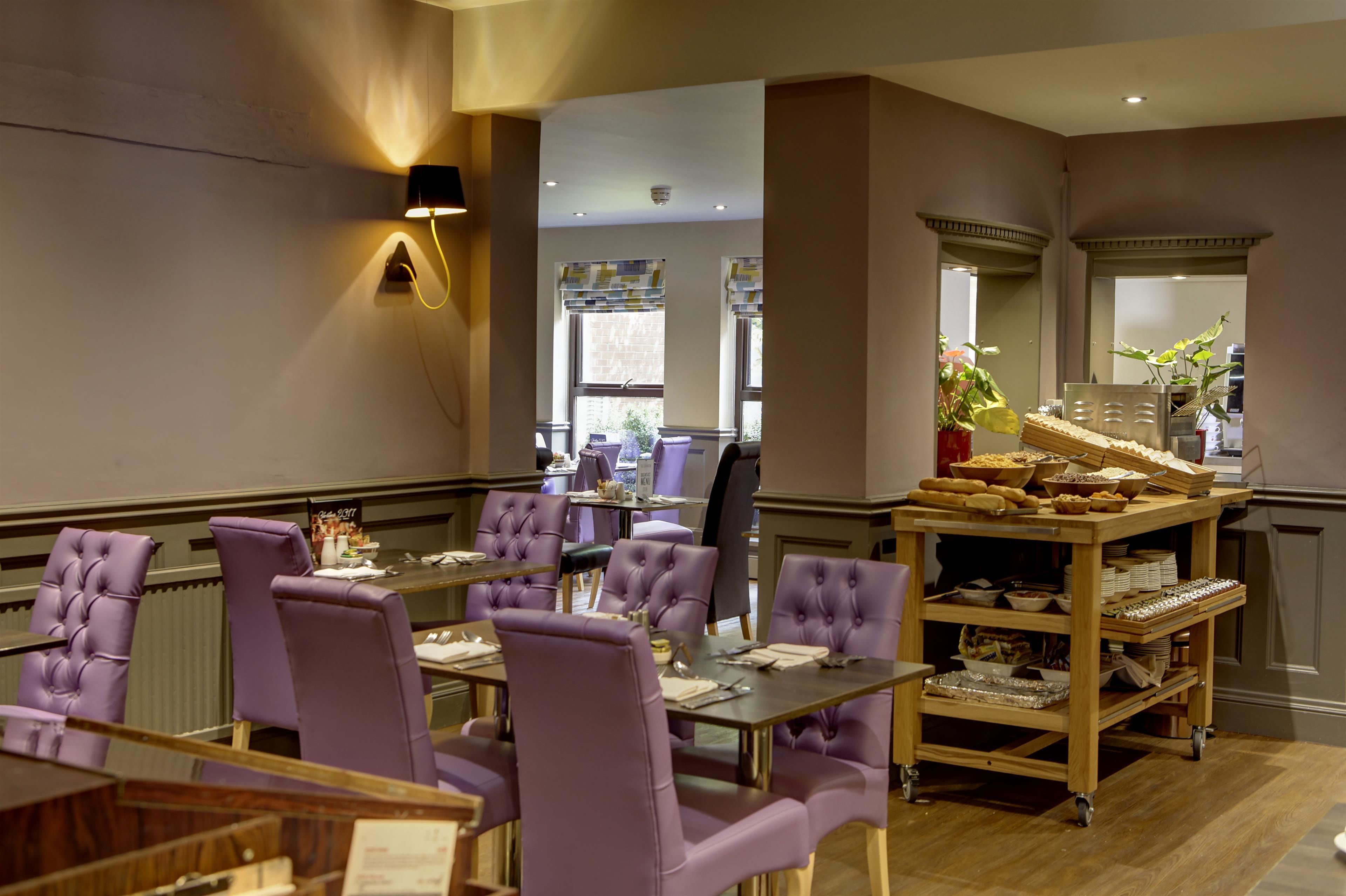 A dining area with purple upholstered chairs and tables, a buffet cart with food items, and a wall-mounted light fixture.