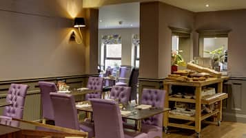 A dining area with purple upholstered chairs and tables, a buffet cart with food items, and a wall-mounted light fixture.