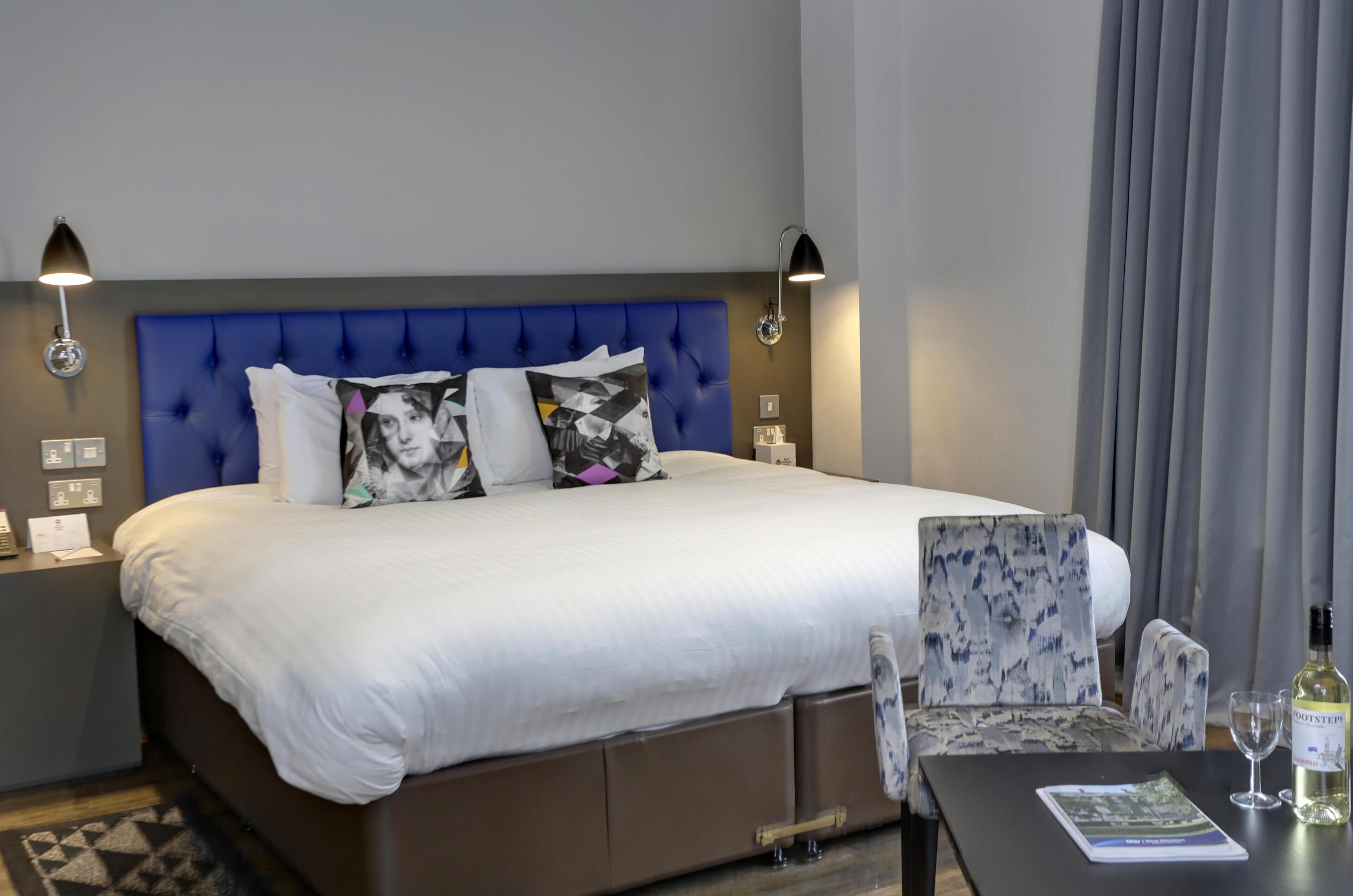 Una habitación de hotel con una cama grande, dos lámparas de noche, una mesita de noche, una silla, una mesa con un vaso y una botella, y una revista.