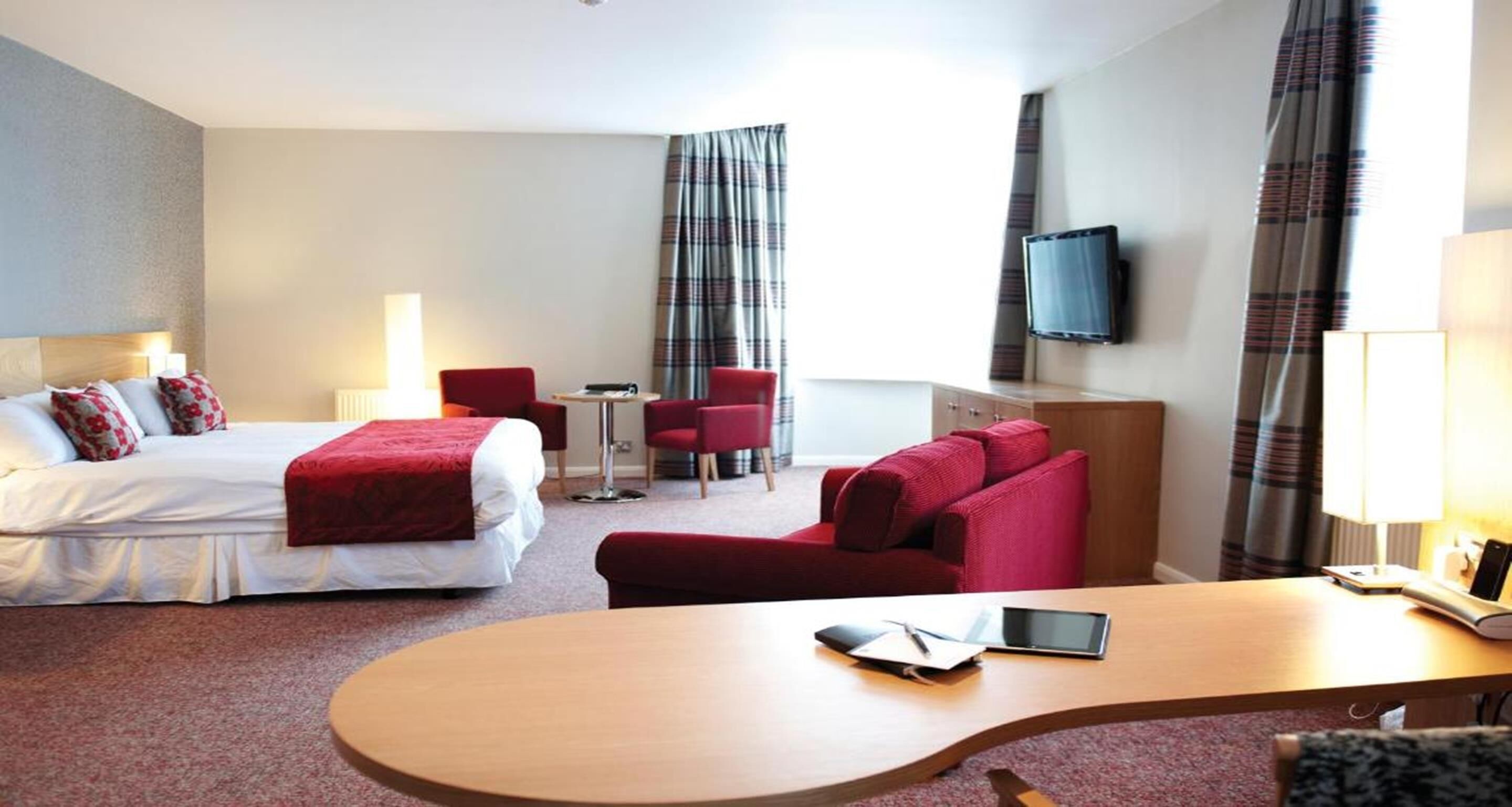 Una habitación de hotel con cama, escritorio, sofá rojo, televisión y lámpara.