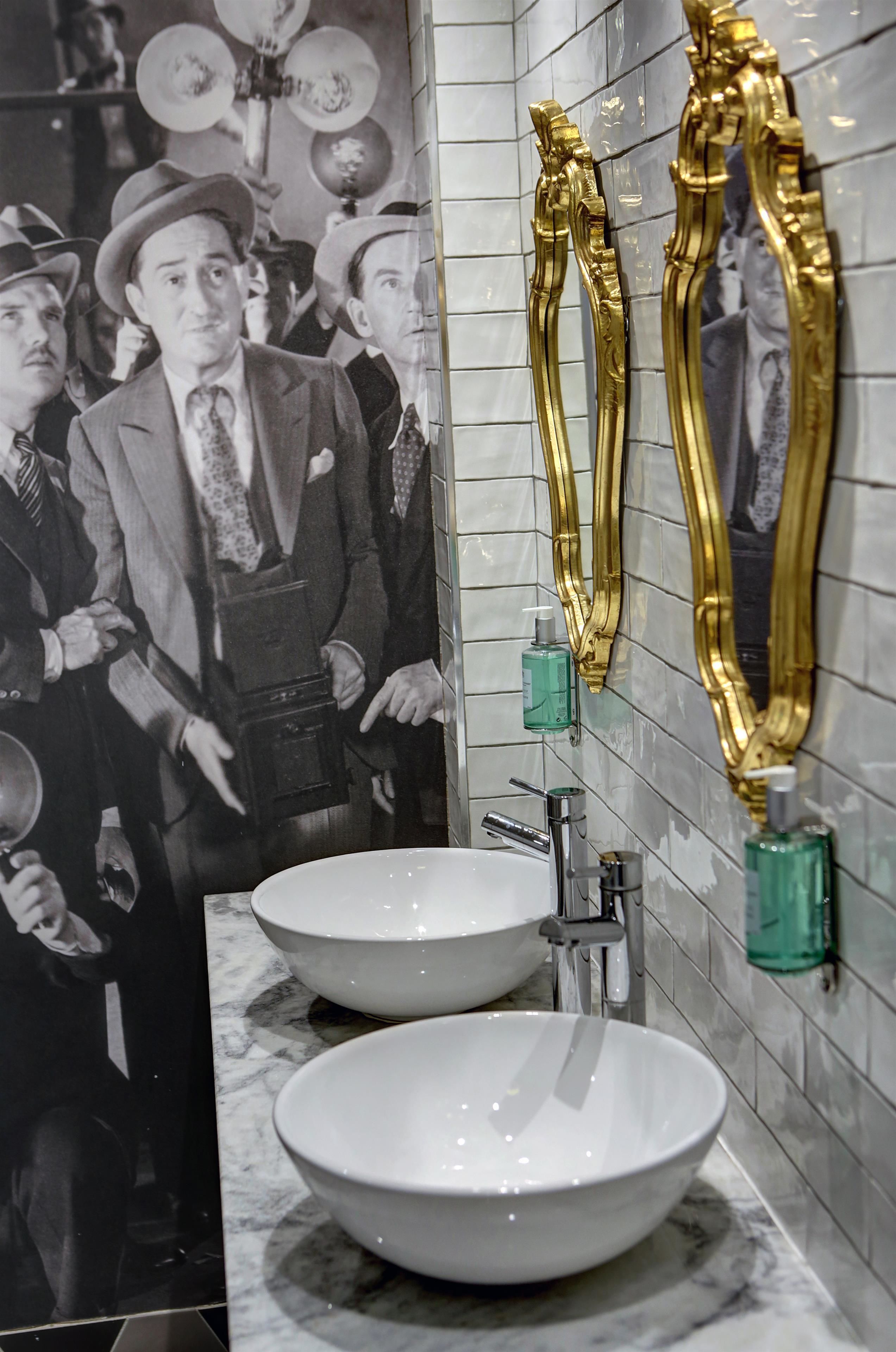 A bathroom with two white sinks, a mirror with gold handles, and a poster of men in suits.
