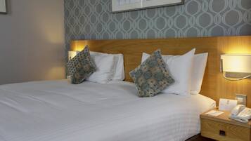 Una habitación de hotel con una cama bien hecha, dos almohadas estampadas, lámparas de noche, un teléfono en la mesita y un cuadro en la pared.