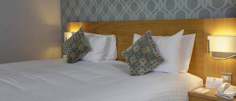 A hotel room with a neatly made bed, two patterned pillows, bedside lamps, a bedside telephone, and a framed picture on the wall.