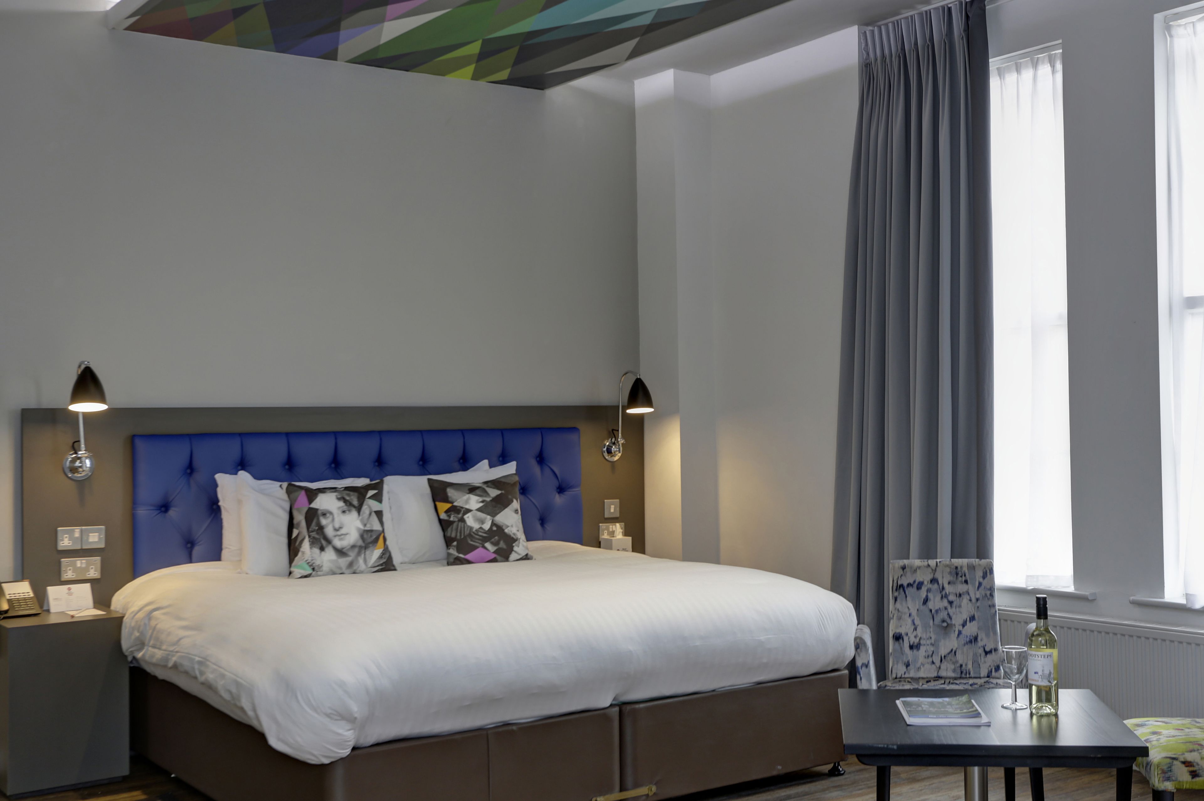A hotel room with a large bed, a bedside table with a bottle and glass, and a view of the outside through a window with curtains.
