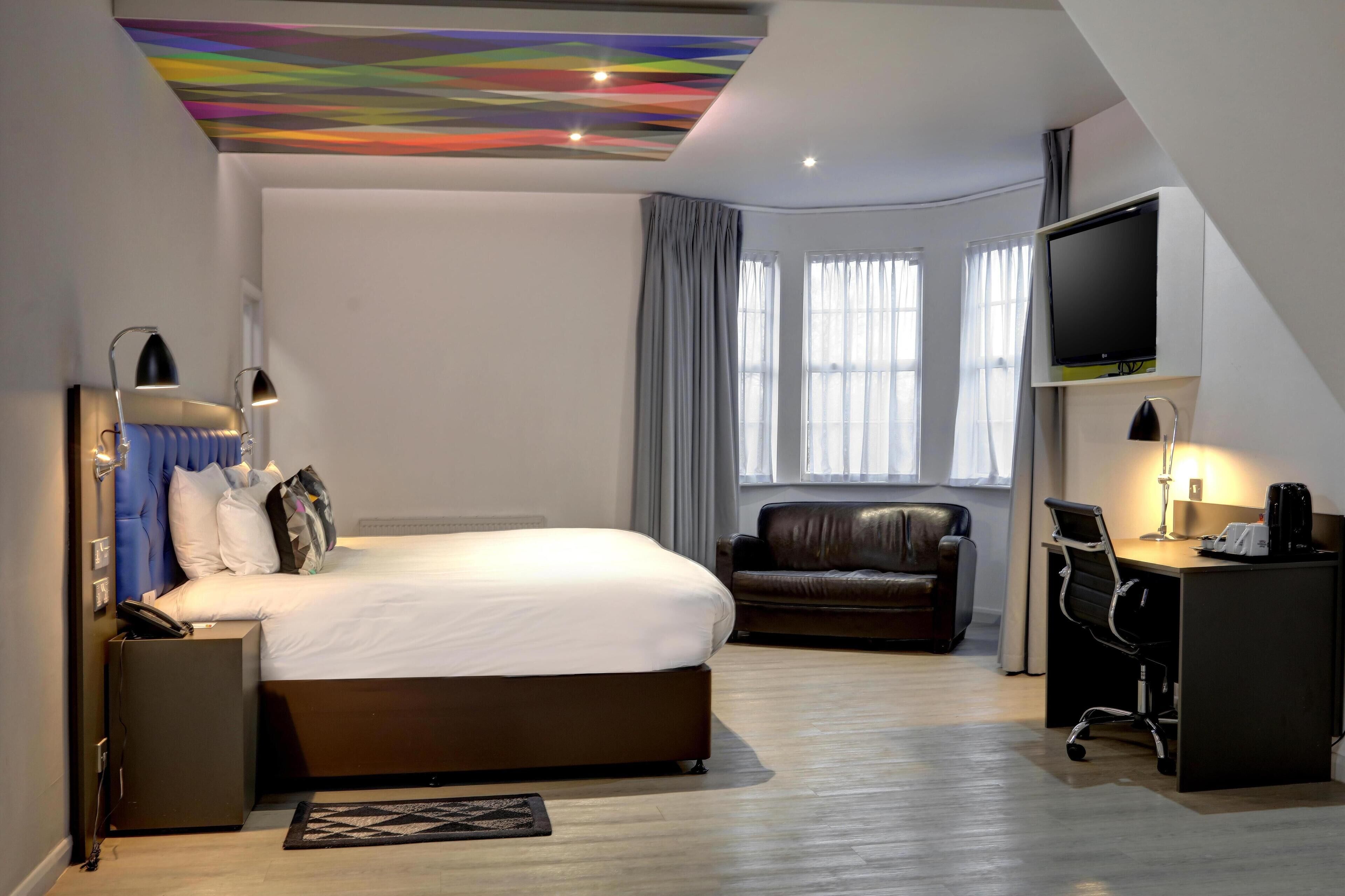 A modern hotel room with a large bed, a leather sofa, a desk with a chair, a television, and a colorful ceiling.
