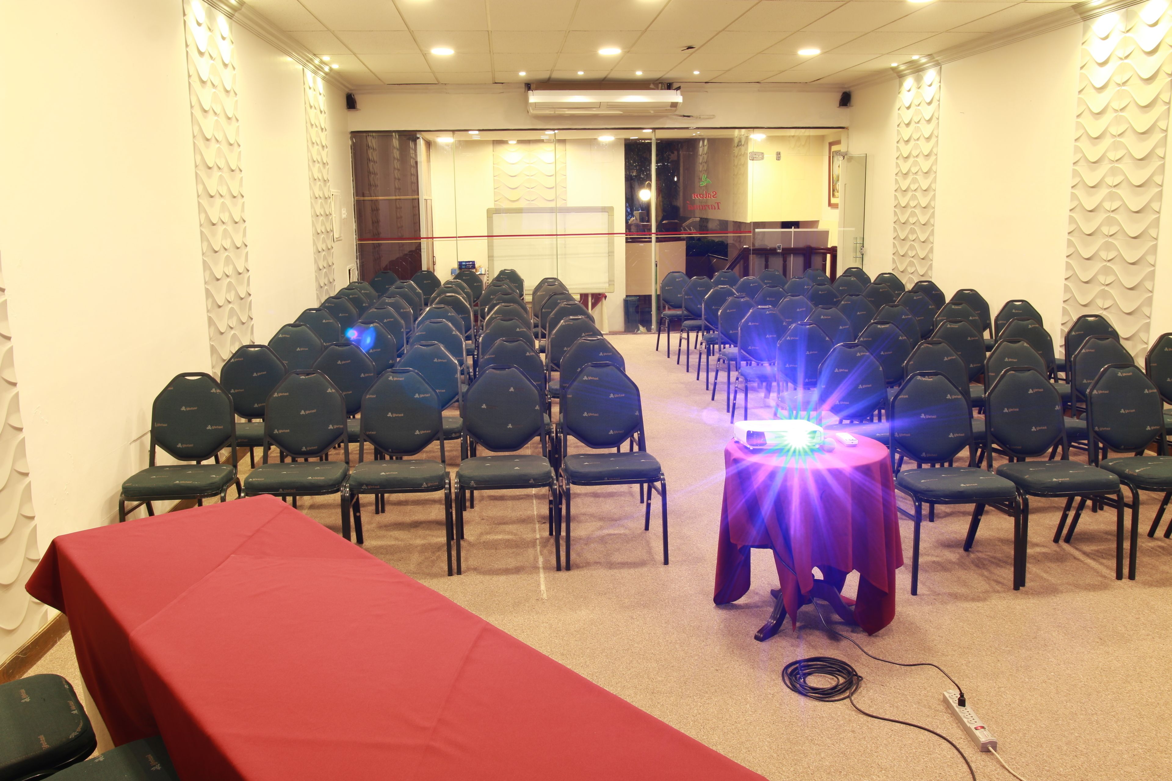 Un salón de conferencias con filas de sillas, una mesa con mantel rojo y un proyector sobre una mesa.