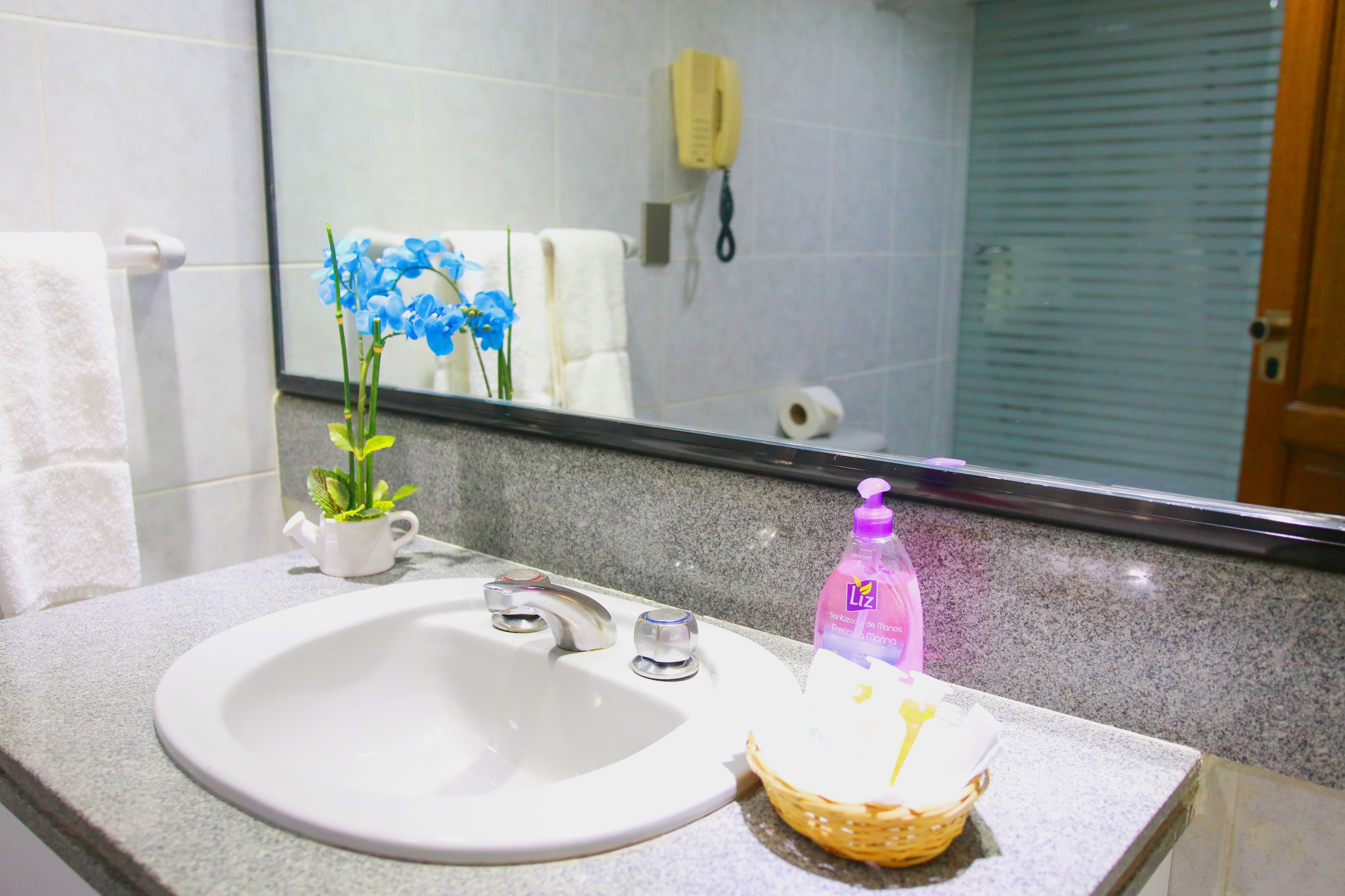 Un lavamanos con un lavabo blanco, una pequeña maceta con flores azules, una botella de jabón para manos y una cesta con un objeto amarillo.