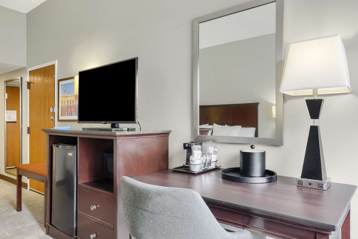 Une chambre d’hôtel équipée d’un bureau en bois, d’une télévision, d’un miroir, d’une lampe, d’un réfrigérateur, d’une machine à café et d’une chaise.