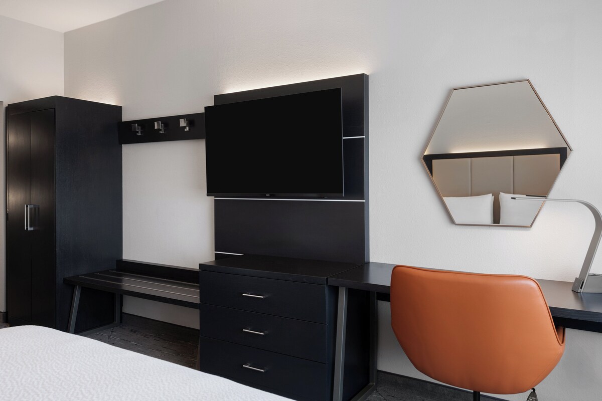 A modern hotel room with a black desk, a flat-screen TV, a large mirror, and a brown chair.