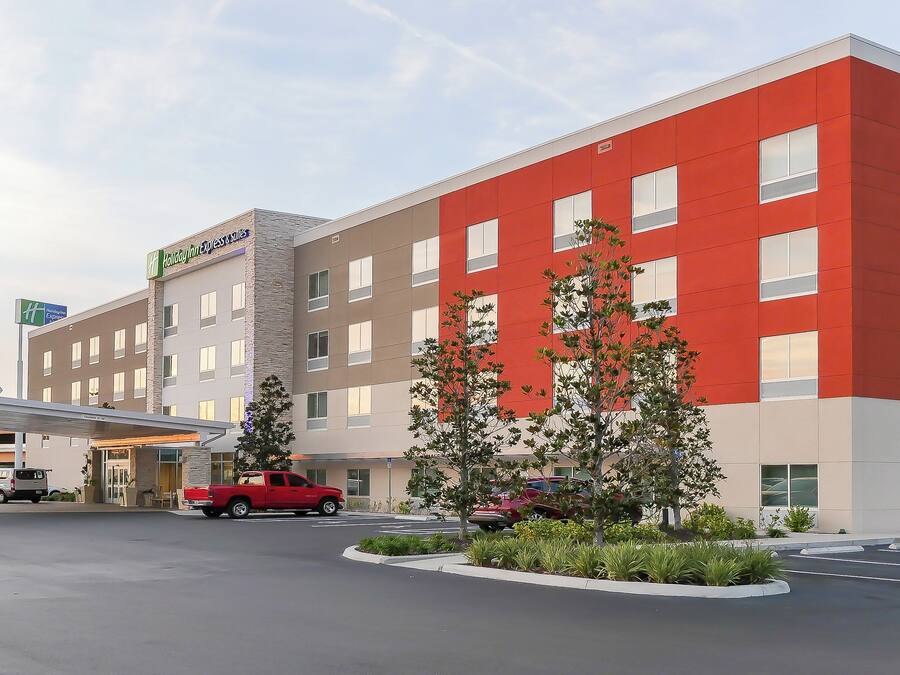 Ein Holiday Inn Express Hotel mit modernem Design, das eine rote und beigefarbene Fassade, einen überdachten Parkbereich und einen begrünten Vorgarten mit Bäumen zeigt.