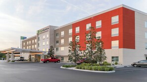 Un hotel Holiday Inn Express con diseño moderno, que presenta una fachada roja y beige, una zona de estacionamiento techada y un jardín delantero arbolado.
