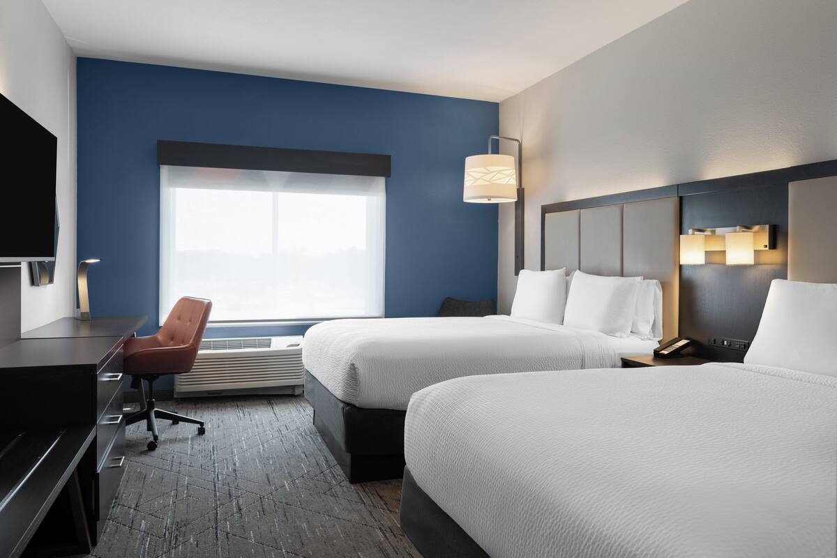 A hotel room with two beds, a desk, a chair, a large window, and a wall-mounted lamp.