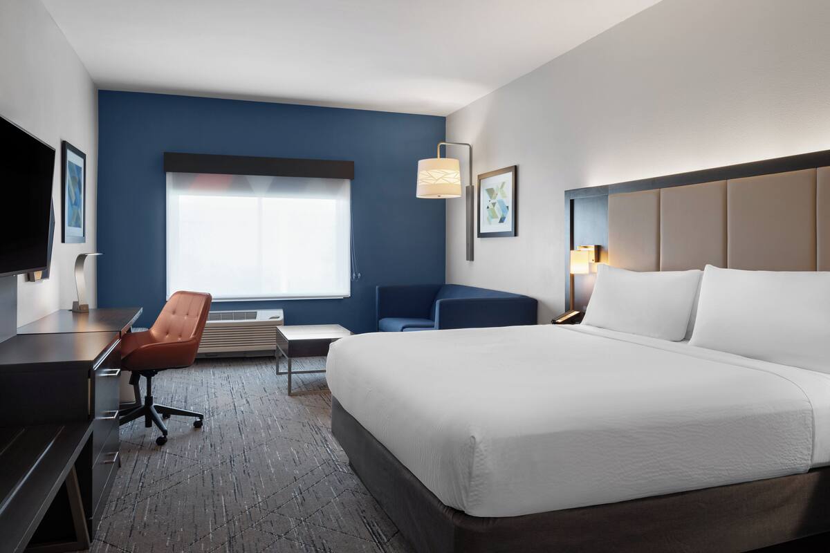 A hotel room with a large bed, a desk, a chair, a TV, and a window with blinds.