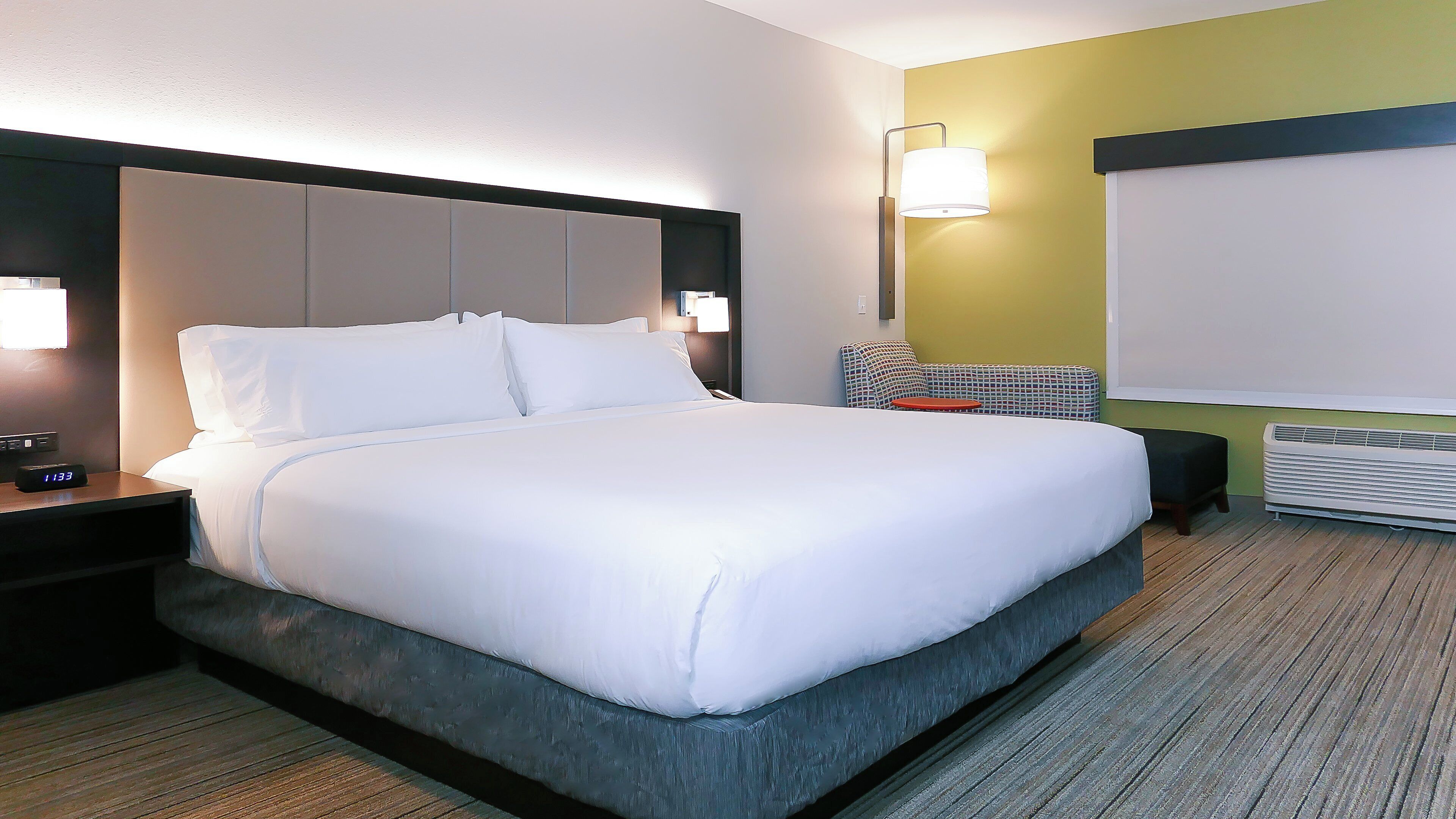 A hotel room with a bed, bedside table, lamp, and a view of the outside.