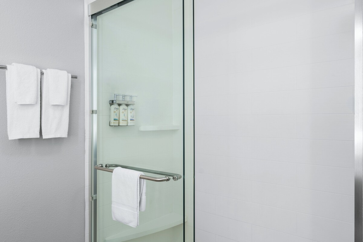 A modern bathroom with a glass shower enclosure, a towel rack with two white towels, and a small shelf with toiletries.