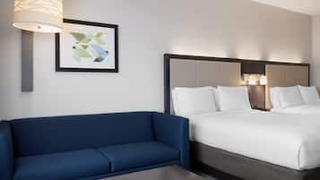 Habitació d'hotel amb un sofà blau, un llit amb roba de llit blanca, una obra d'art abstracta emmarcada i una làmpada penjada a la paret.