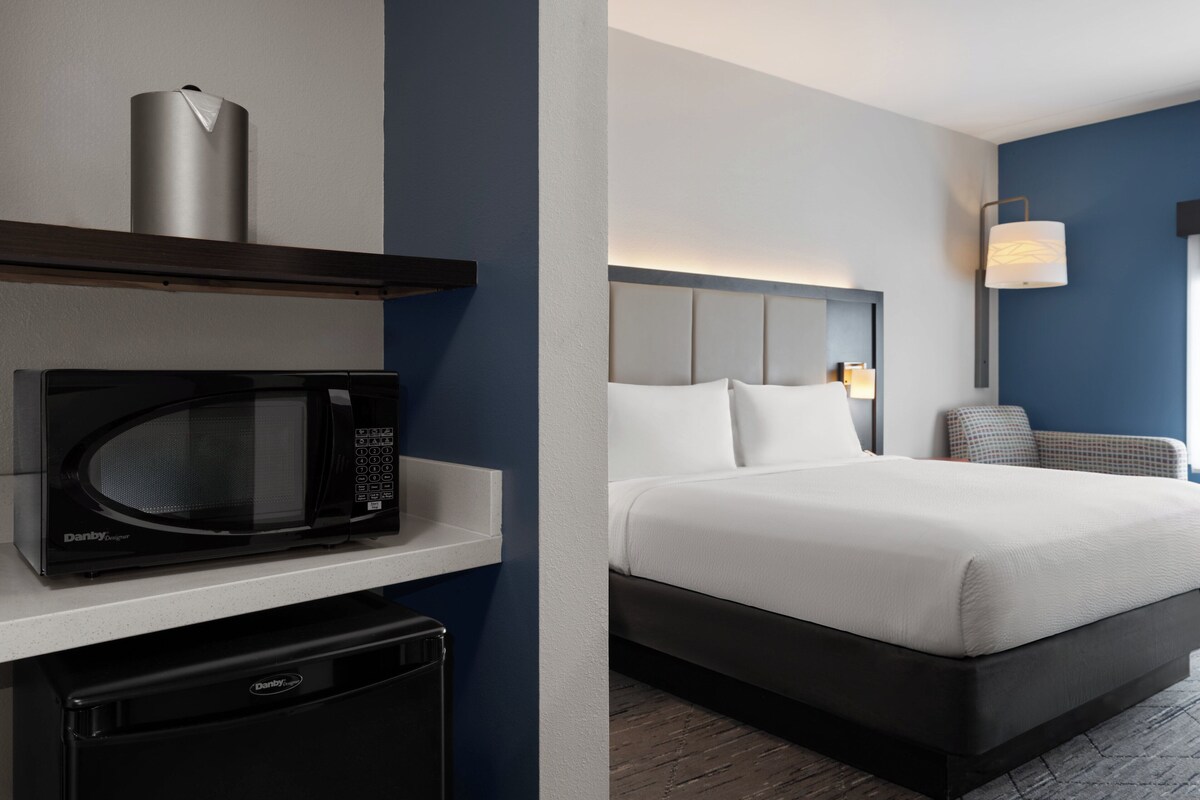 A hotel room with a bed, a microwave, a mini fridge, a nightstand, and a wall-mounted lamp.