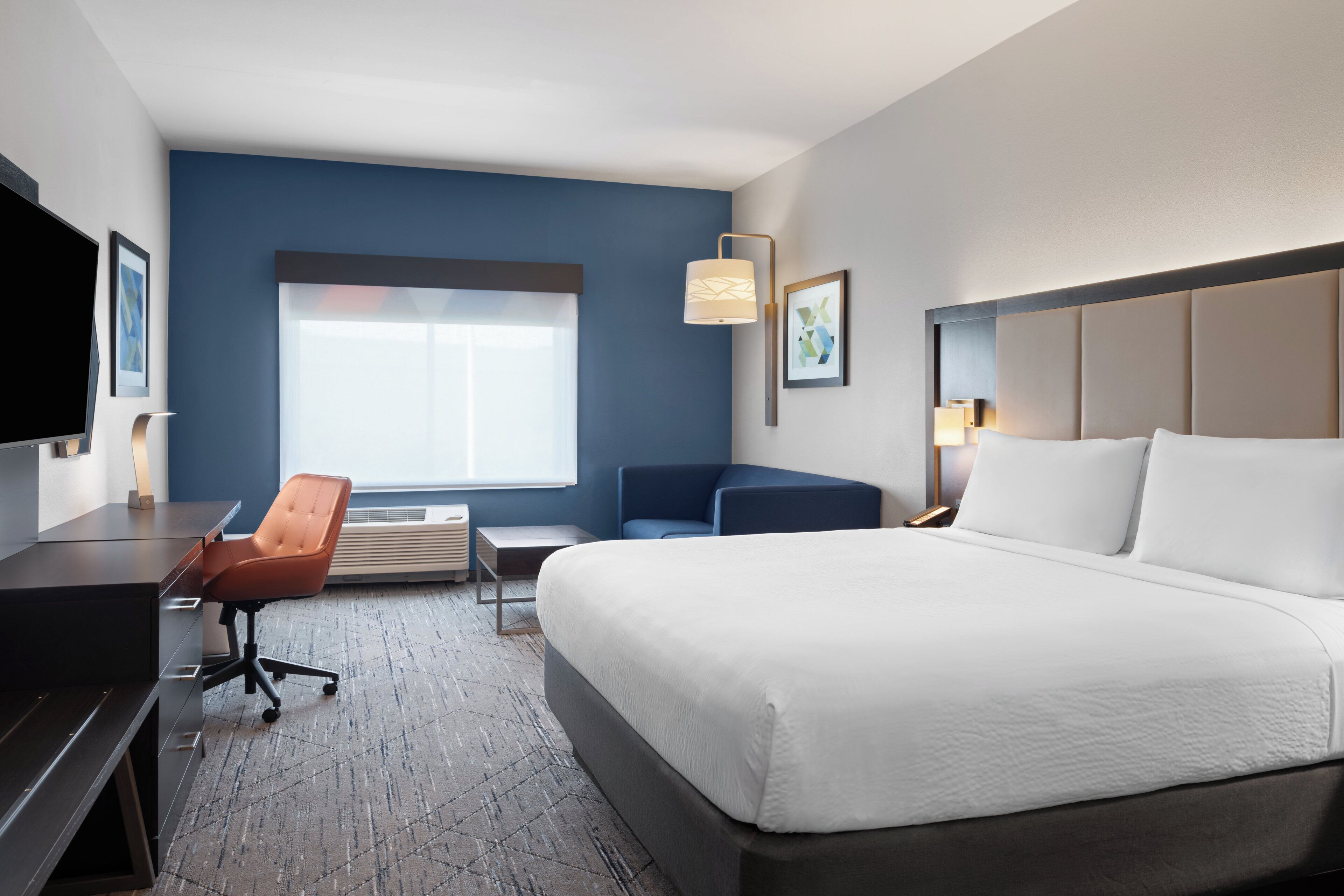 Habitación de hotel con cama, escritorio, silla, televisor, ventana y lámpara de pared.