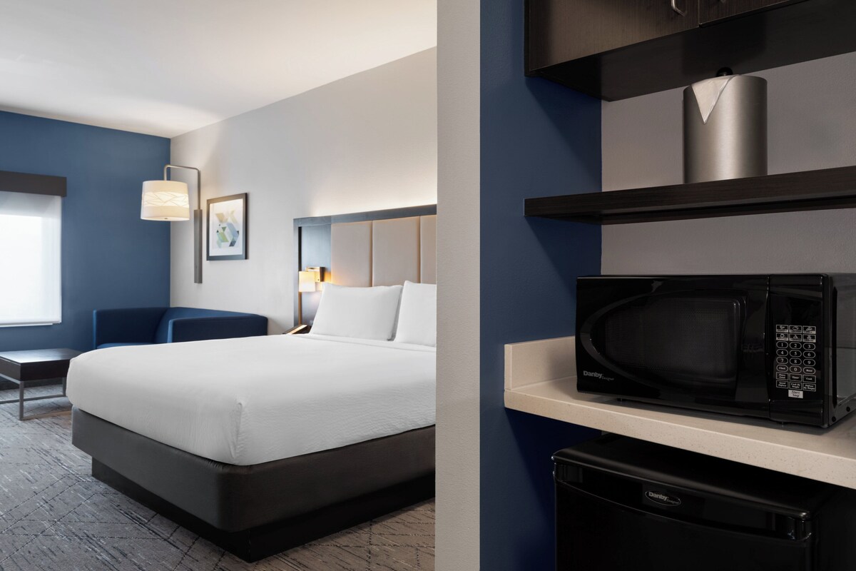 A hotel room with a bed, a desk, a chair, a microwave, and a shelf with a container.