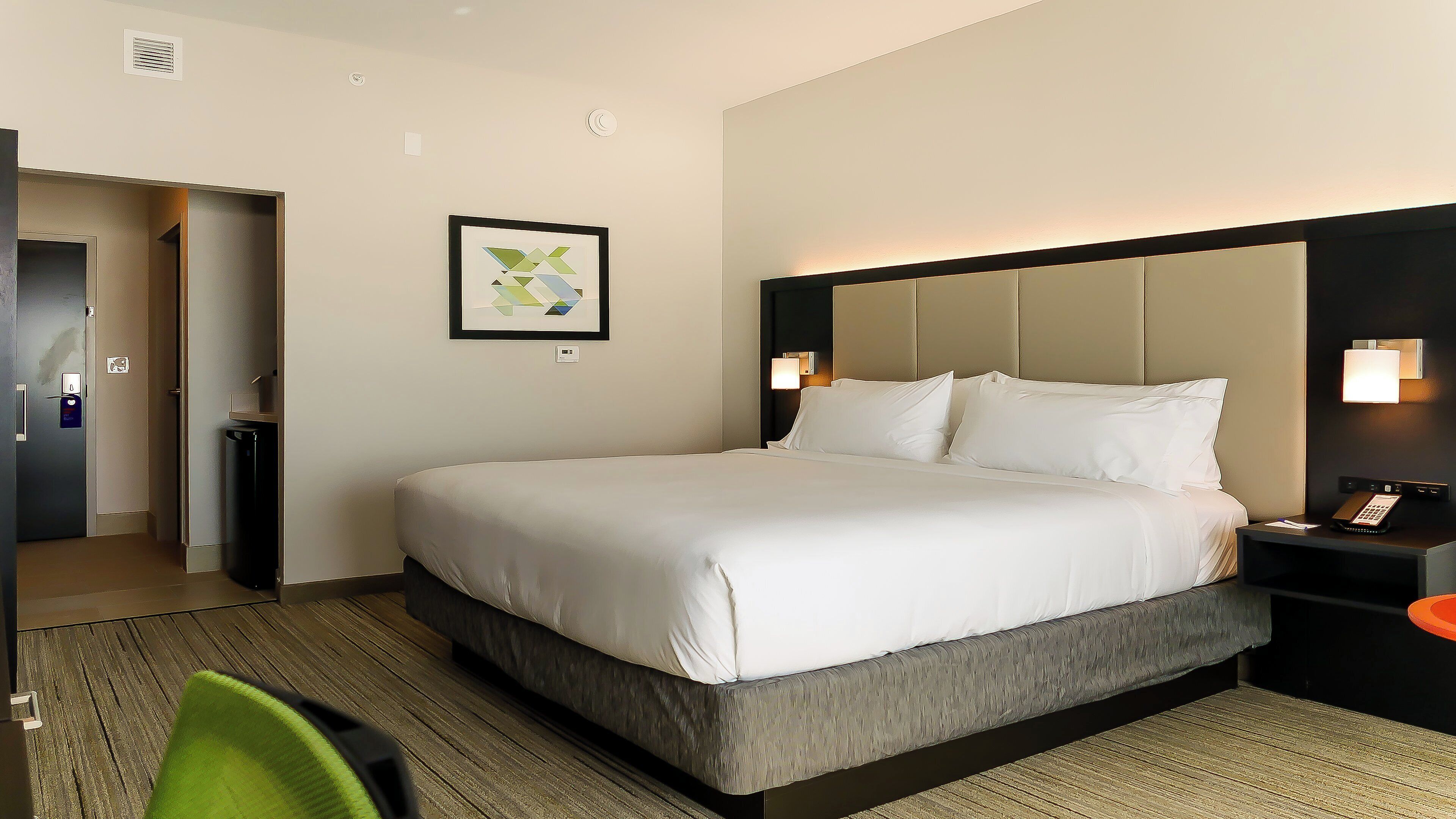 A hotel room with a large bed, a bedside table with a lamp, a wall-mounted artwork, and a door leading to a bathroom.