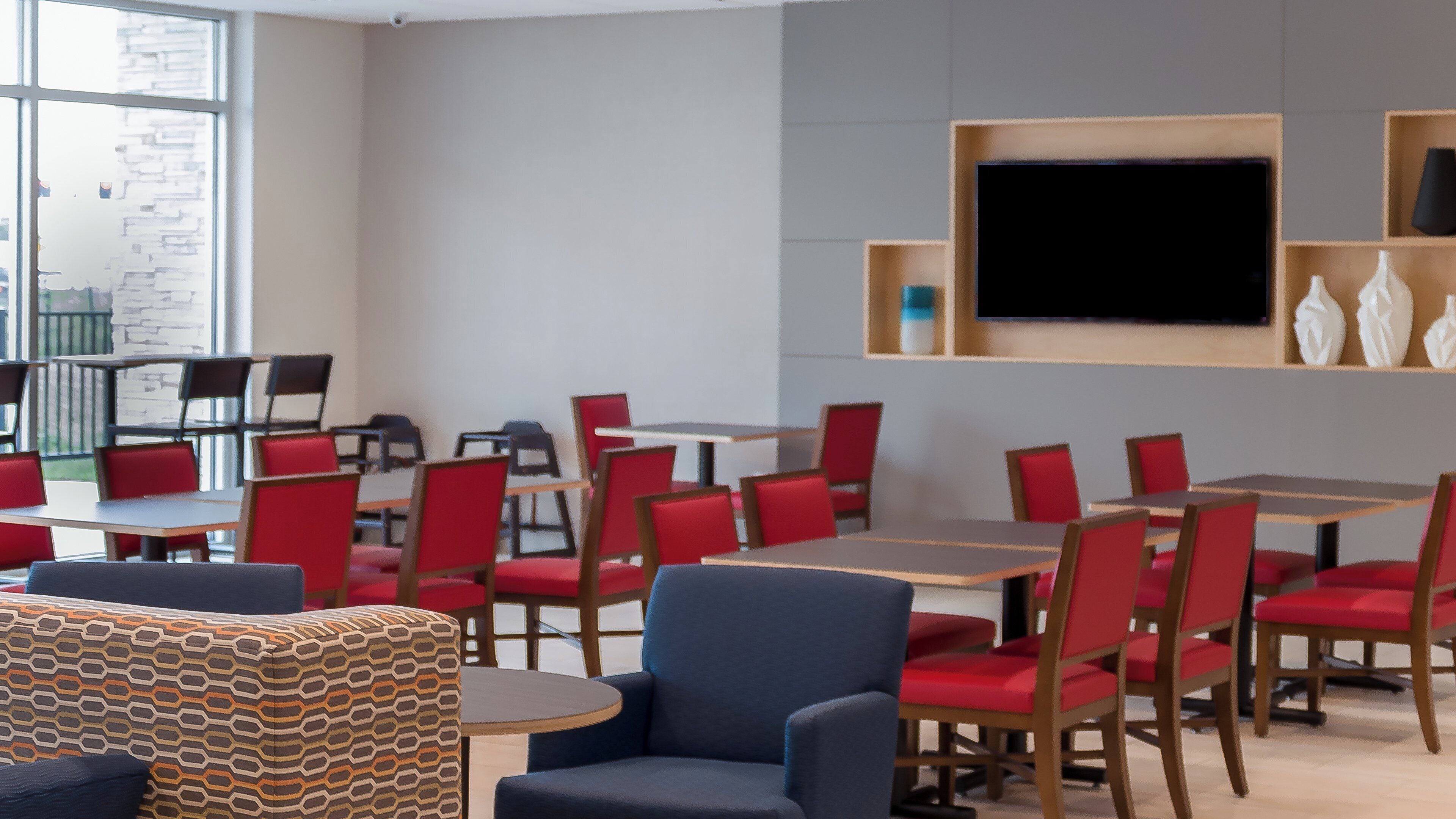 A modern dining area with red chairs and tables, a large flat-screen TV mounted on the wall, and a patterned ottoman in the foreground.
