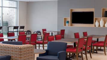 A modern dining area with red chairs and tables, a large flat-screen TV mounted on the wall, and a patterned ottoman in the foreground.
